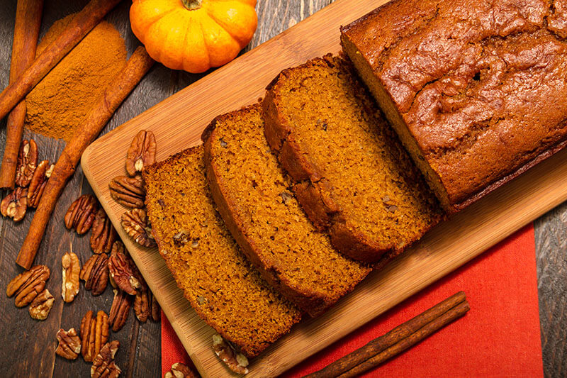 Fresh baked loaf of pumpkin bread sitting on wood cutting board with pecan nuts, spices and orange napkin